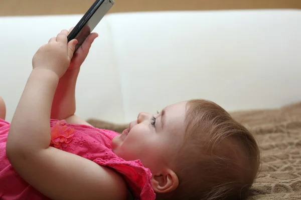 Pequeña linda chica acostada en el sofá y jugando con un teléfono inteligente — Foto de Stock