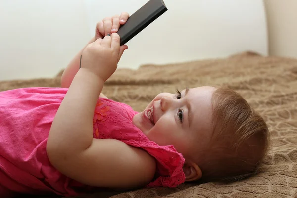 Lilla leende barn leker med din smartphone liggande på en soffa — Stockfoto