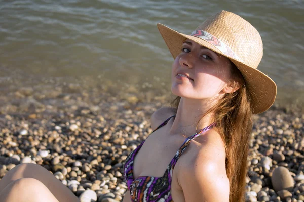 Beautiful young woman with sun hat sitting and smiling on the beach. Sea in background. horizontal — Stock Photo, Image