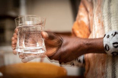 Double exposure that creates a blurry effect on the foreground of an African lady's hands as she tries to steadily hold a glass. Concept of difficulties due to the tremors of Parkinson's disease