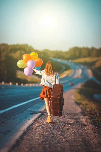 Happy young woman with balloons and large suitcase walking along road. Freedom, summer travel