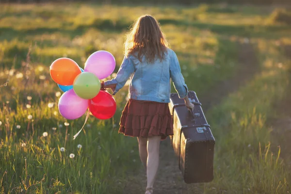 漂亮的女孩与大皮箱和彩色的气球领域的路上走。阳光，日落，野花，春天。享受自由和旅行 图库图片
