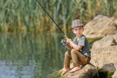 Küçük çocuk taşın üzerinde oturuyor ve balık tutuyor.