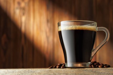 Işık demeti şeffaf bir fincan espresso üzerine düşer, kahve çekirdekleri etrafa saçılmış, ahşap arka planda..