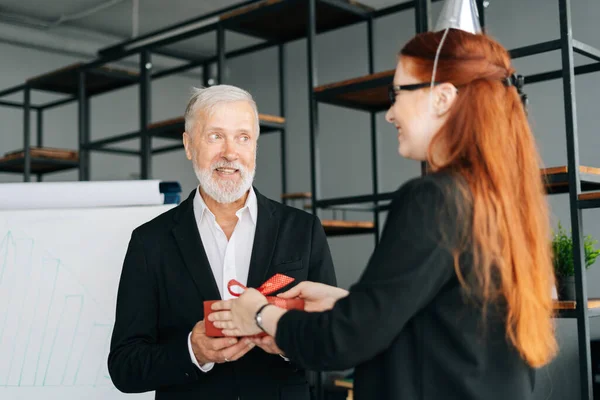 Business woman giving red gift box to smiling mature adult male leader during the Happy Birthday