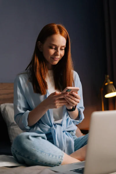 Portrait of charming redhead young woman typing message on mobile phone at apartment.