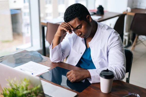 Tired African black male doctor working on laptop, thinking about problem and drinking coffee.