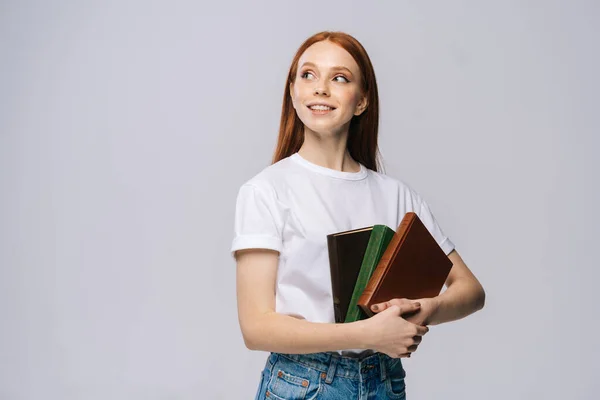 Smiling young woman college student holding books and dreamy looking away on isolated background.