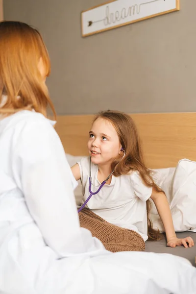 Back view of happy little girl listening heartbeat with stethoscope to female doctor sitting in bed.