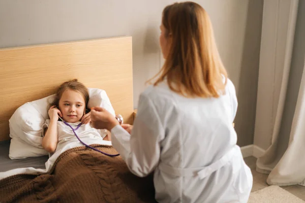 Happy little girl listening heartbeat with stethoscope to female doctor sitting in bed.