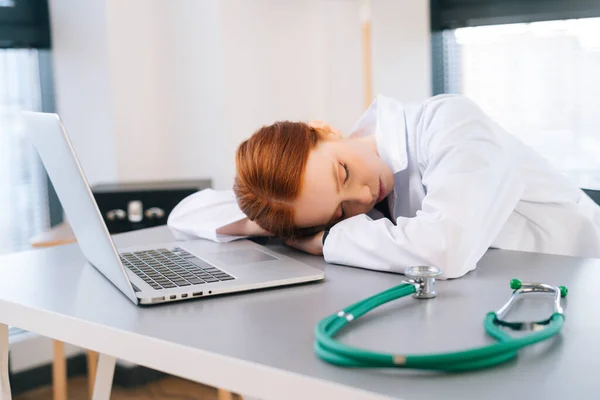 Close-up face of tired exhausted overworked young female doctor in white coat sleeping lying at desk with laptop and stethoscope.