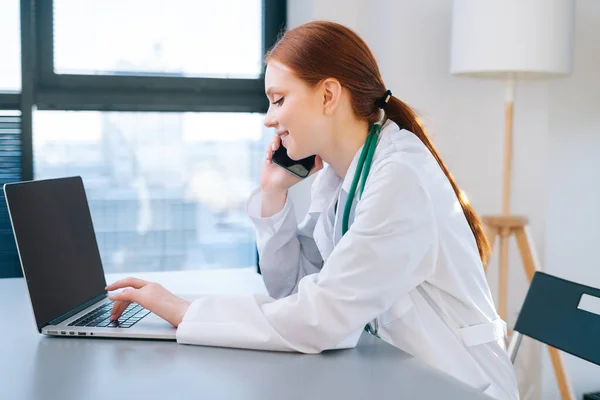 Side view of happy smiling female doctor in white coat typing on laptop keyboard and talking on mobile phone with patient during remote consultation.