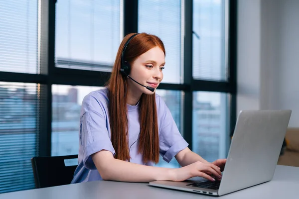 Close-up of focused young woman operator using headset and laptop during customer support at home office.
