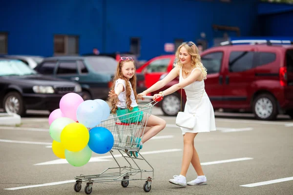 Kleines Kind und Schwester mit Sonnenbrille vergnügen sich draußen mit vielen bunten Luftballons. Kind sitzt im Einkaufswagen in der Nähe von Einkaufszentrum. glücklich lächelndes Kind im Warenkorb. — Stockfoto