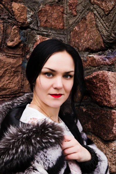 Young fashion elegant woman in white dress and fur coat at the brick wall — Stock Photo, Image