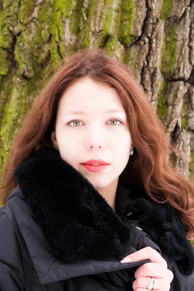 Young woman wearing warm coat, standing against tree's bark back — Stock Photo, Image