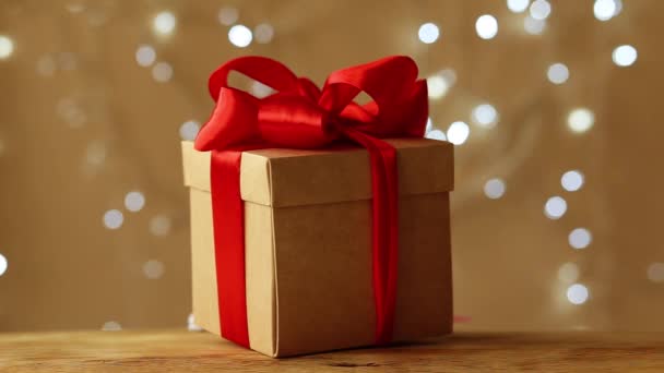 Pudełko zapakowane w czerwoną wstążkę stoi brązowe tło. Girlanda świąteczna błyszczy w tle — Wideo stockowe