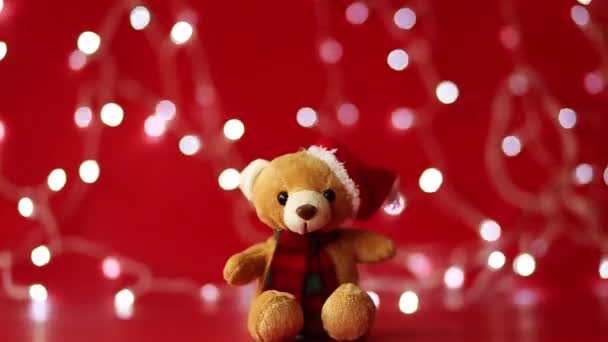 Um urso de pelúcia de brinquedo macio em um chapéu e cachecol se senta em um fundo vermelho e uma grinalda de Natal piscando no fundo — Vídeo de Stock