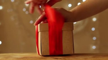 Kadınların elleri hediyeyi bir kutuya koyup bej arka planda kırmızı bir kurdele ve parlak bir çelenkle bağlıyorlar.