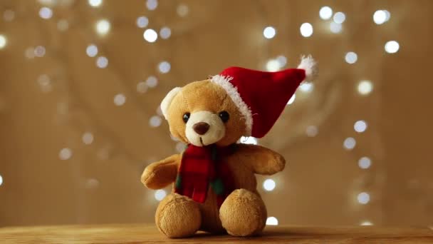 Un osito de peluche de juguete suave en un sombrero y una bufanda se sienta sobre un fondo beige y una guirnalda de Navidad parpadeante en el fondo — Vídeos de Stock