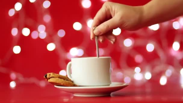 Uma xícara branca com um pires e um biscoito fica contra um fundo de vermelho e uma grinalda piscando. A mão feminina interfere com uma colher em um copo — Vídeo de Stock