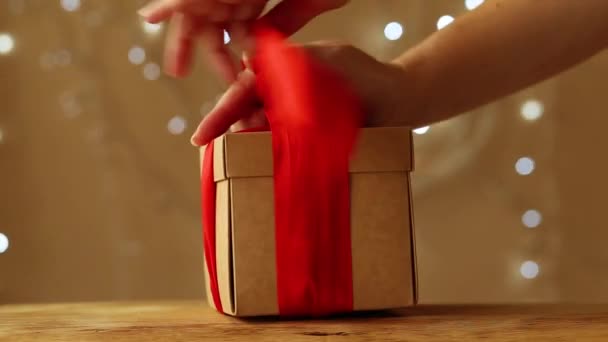 Женские руки заворачивают подарок в коробку и связывают красной лентой на бежевом фоне и сверкающей гирляндой — стоковое видео