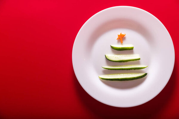 Новогодний огурец в виде елки на белой тарелке на красном фоне. Вид сверху. Принято. Горизонтальное фото