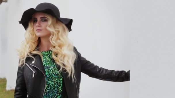 Piękna blondynka w czarnym kapeluszu i czarnej skórzanej kurtce odwraca się do kamery i pokazuje swój język — Wideo stockowe