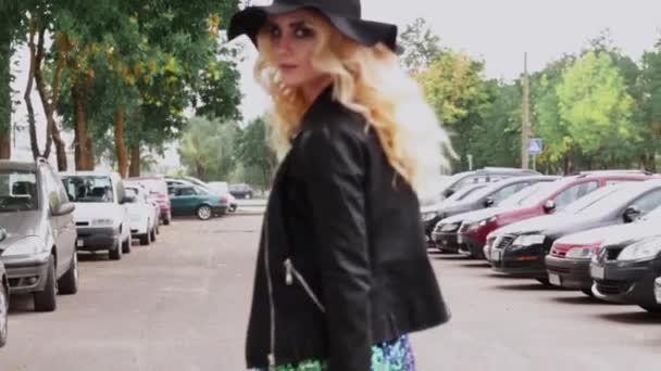 A stylish blonde girl in a black hat, black leather jacket and a shiny dress walks along the parking lot along the cars and turns around — Stock Video