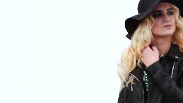Elegante ragazza bionda in un cappello nero, giacca di pelle nera e un abito lucido in posa davanti alla fotocamera su uno sfondo bianco muro — Video Stock
