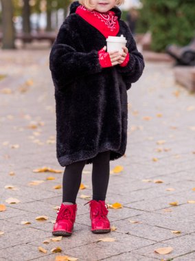 Sonbahar havasında elinde bir bardak sıcak çikolatayla şehir caddesinde poz veren siyah ceketli, kırmızı kaşkollü ve botlu bir kız. Fotoğraf.
