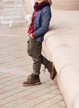 Pantolon giymiş şık bir çocuk, bir ceket ve elinde bir bardak sıcak içecekle şehir caddesinde poz veren bir eşarp. Fotoğraf.