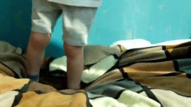 Açık renkli tişörtlü, şortlu ve çoraplı küçük bir çocuk kareli yatak örtüsüyle kaplı bir yatakta zıplıyor. Odada çok güneş ışığı var. Yüksek kaliteli FullHD görüntüler