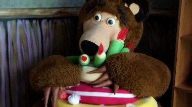 Kahverengi ayı boyutunda bir kukla, elinde büyük bir oyuncak pastayla rahat bir koltukta oturur. Ayı pastayı masaya koyar ve tok olduğunu gösterir. Yüksek kaliteli FullHD görüntüler. Minsk, Belarus - 5 Eylül 2021