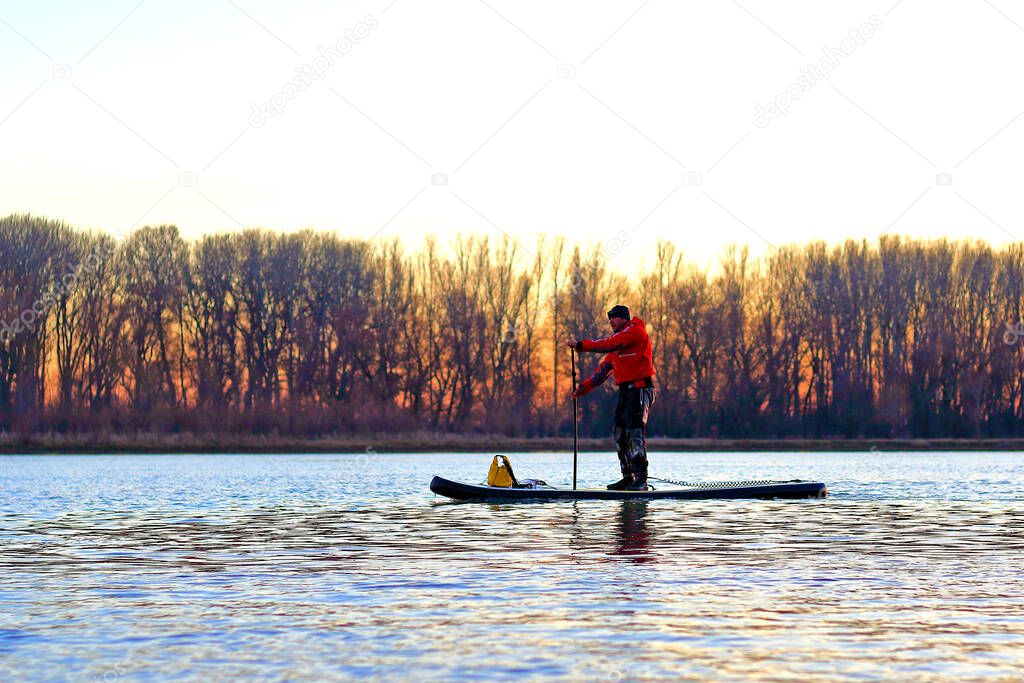 Athletic man standing with a paddle on the stand up paddle board (paddleboard, SUP) in the winter Danube river against of trees at sunset