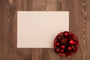 Minimalist Noel dairesi boş bir kağıt tabaka ve ahşap arka planda kırmızı parıltı ve süs eşyalarıyla dolu bir sepet ile yatıyordu. Bayram kutlamaları, davetiyeler veya mevsimlik tasarımlar için mükemmel..