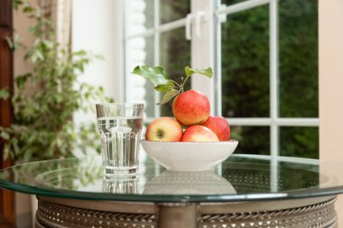 Cam bir masada bir bardak su ve bir kase kırmızı elma. Sağlıklı bir yaşam tarzı için doğal detoks, hidrasyon ve günlük vitamin besinlerinin sembolü..