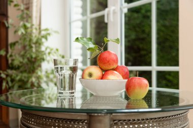 Hapların yanında bir kasede taze kırmızı elmalar ve cam bir masanın üzerinde bir bardak su. Doğal beslenme ve tıbbi takviyeler arasındaki dengenin sembolü. Sağlık, dokunulmazlık ve refah kavramı