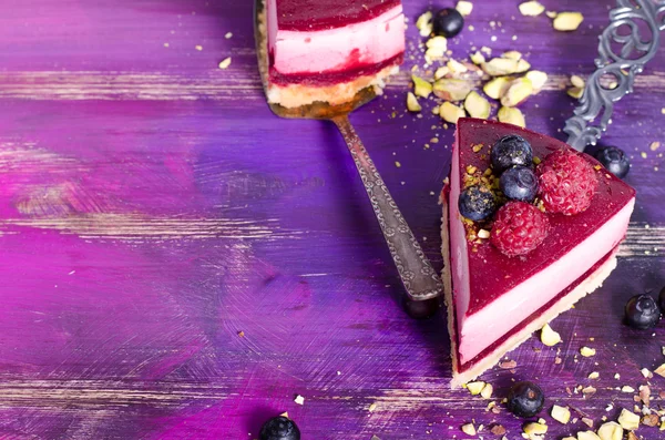 Piece of delicious raspberry cake with fresh raspberries, blueberry, currants and pistachios on shovel, bright purple, violet background. Free space for your text. — Stock Photo, Image