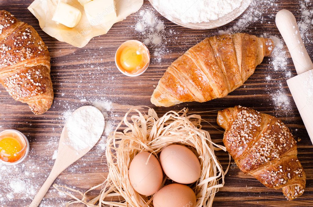 Ingredients for baking croissants - paper, flour, wooden spoon, rolling pin, eggs, egg yolks, butter served on a rustic wooden tray table. 