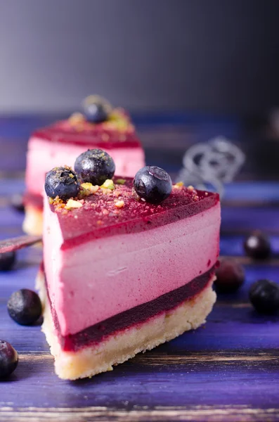 Piece of delicious raspberry cake with fresh raspberries, blueberry, currants and pistachios on shovel, bright purple, violet, blue background. Free space for your text. — Stock Photo, Image