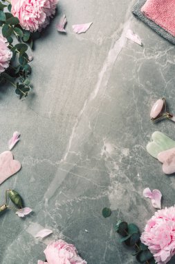 Yüz silindiri, gua sha masaj aleti, yağ şişesi, havlu, mermer arka planda pembe şakayıklar. Uzayı kopyala Düz yatıyordu. Üst Manzara. Rutin cilt bakımı, vücut bakımı, spa konsepti
