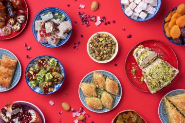 Kırmızı arka planda Orta Doğu tatlıları. Arap tatlısı, baklava, halva, rahat lokum, şerbet, fındık, şam fıstığı, hurma, kuru üzüm, kayısı, kilise kilisesi, kurabiye. Üst Manzara. Düz yatıyordu. Boşluğu kopyala.