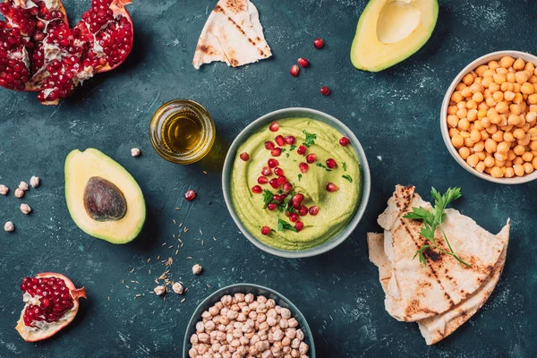 Homemade green hummus with avocado, parsley, olive oil, pita, raw chickpeas, pomegranate on dark background. Middle eastern, jewish, arabic cuisine. Top view. Copy space. Vegan, vegetarian food.