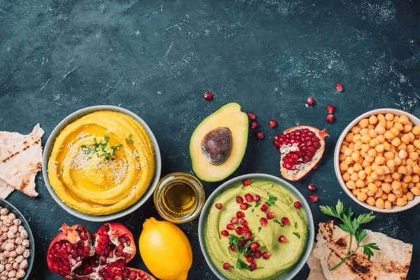 Ingredients for yellow hummus and green hummus - chickpea, tahini, olive oil, sesame seeds, pita, avocado, pomegranate on dark background. Middle eastern, jewish, arabic cuisine. Top view. Copy space.