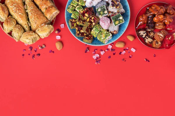Kırmızı arka planda Orta Doğu tatlıları. Arap tatlısı, baklava, halva, rahat lokum, şerbet, fındık, şam fıstığı, hurma, kuru üzüm, kayısı, kilise kilisesi, kurabiye. Üst Manzara. Düz yatıyordu. Boşluğu kopyala.