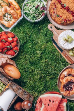 Yeşil arka planda kek, meyve, peynir, şarap ve atıştırmalıklarla yaz pikniği. Üst Manzara. Uzayı kopyala Yaz aile yemeği. Romantik piknik. Sağlıklı yiyecek ve aksesuarlarla piknik sepeti.