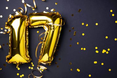 Altın folyo balon numarası, rakam 17. Doğum günü tebrik kartı, 17. yazı. Yıldönümü kutlaması. Pankart. Şık altın rakamlı, açık renkli, parlak, siyah arkaplan. Sayısal rakam.