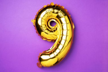 Creative layout. Golden foil balloon number and digit nine 9. Birthday greeting card. Anniversary concept. Top view. Copy space. Stylish gold numeral over purple background. Numerical digit