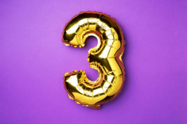 Creative layout. Golden foil balloon number and digit three 3. Birthday greeting card. Anniversary concept. Top view. Copy space. Stylish gold numeral over purple background. Numerical digit